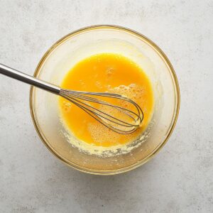 Glass bowl with yellow wet muffin mixture including melted butter, sugar, oil, and egg being whisked on a bright kitchen surface.