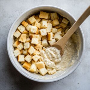 Cubed bread being folded into pineapple stuffing batter with a wooden spoon in a white bowl