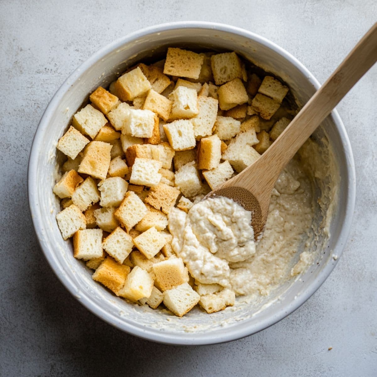 Cubed bread being folded into pineapple stuffing batter with a wooden spoon in a white bowl