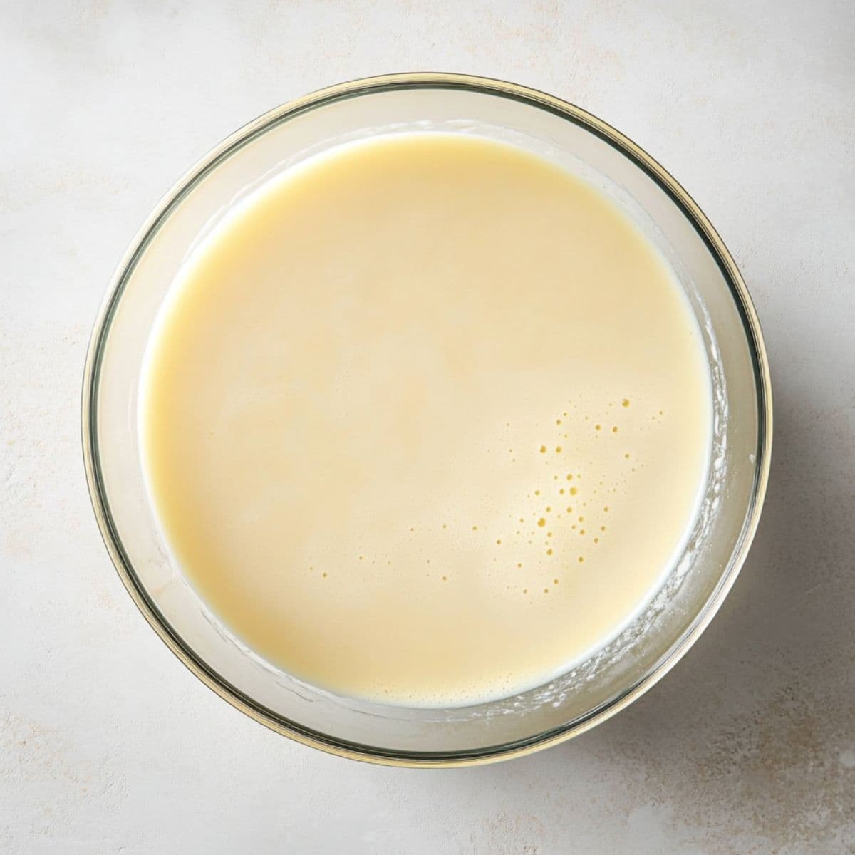 Top-down photo of smooth pale yellow batter in a glass bowl, made from sour cream, milk, and vanilla, ready to be combined with dry ingredients.