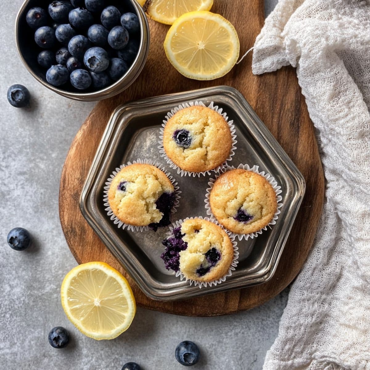 Freshly baked mini blueberry muffins on a metal plate with blueberries and lemon slices styled on a wooden board over a gray surface.