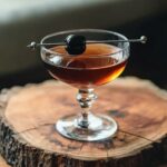 Black Manhattan cocktail in a coupe glass garnished with a cherry, on a wooden surface