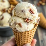 Close-up of butter pecan ice cream scoops in a waffle cone, with visible roasted pecans and creamy vanilla texture.