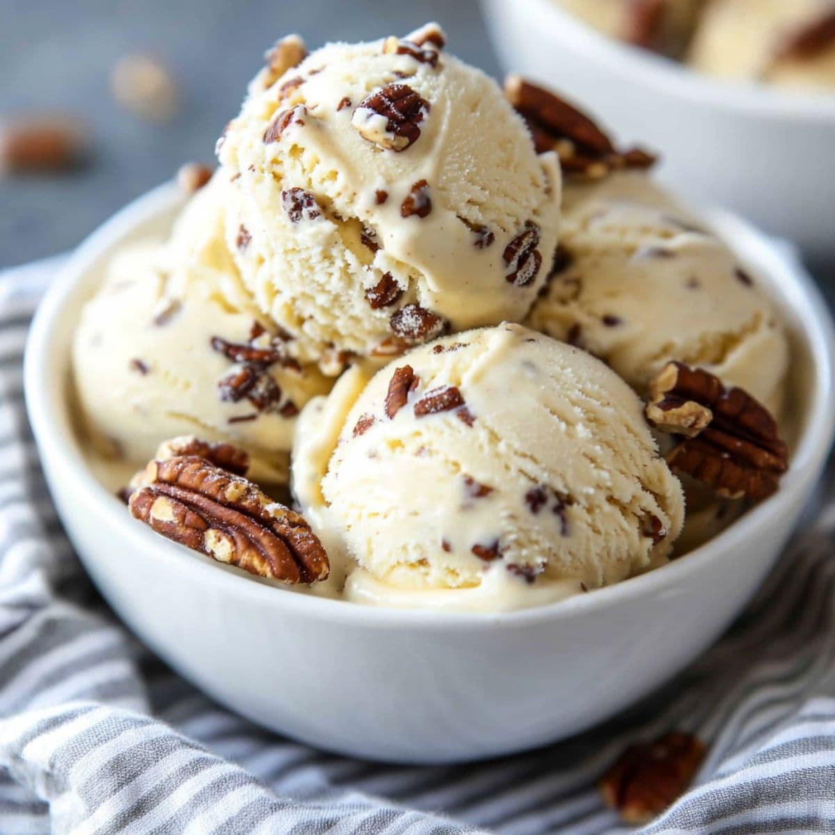 Creamy scoops of butter pecan ice cream served in a white bowl, topped with toasted pecans, on a striped cloth.