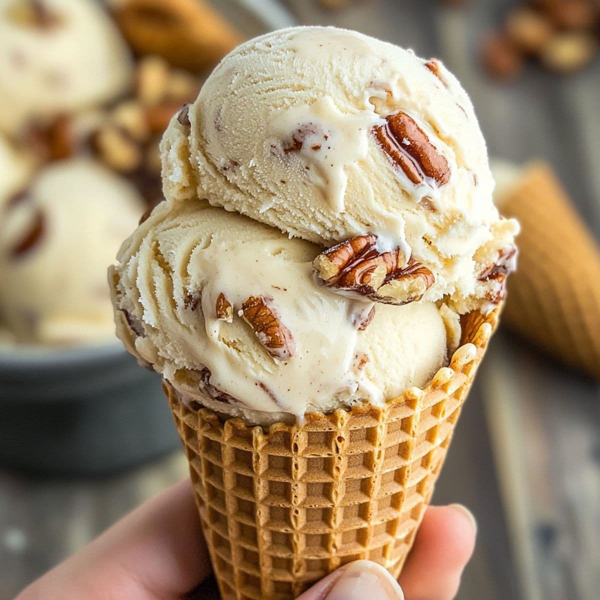 Close-up of butter pecan ice cream scoops in a waffle cone, with visible roasted pecans and creamy vanilla texture.