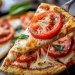 A close-up of a pizza slice topped with fresh tomato, basil, and melted cheese, with a crispy crust, being lifted by a pizza server.