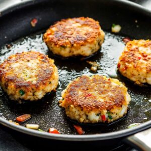 Golden crab cakes cooking in a skillet with olive oil, crisp edges and bits of herbs and bell peppers visible