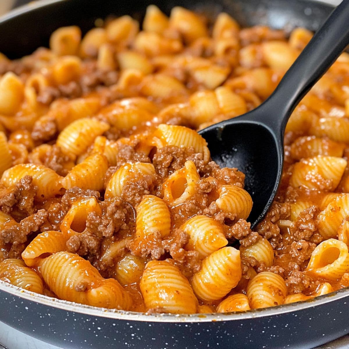Close-up view of creamy beef and shells in a skillet with a black spoon, ready for serving or storage.