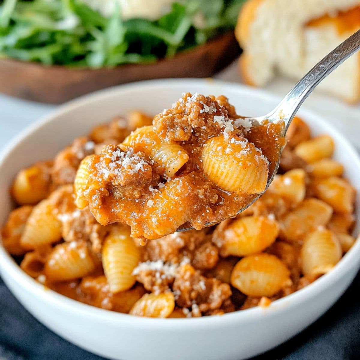 Spoonful of creamy beef and shells lifted over a bowl, with salad and bread in the background.
