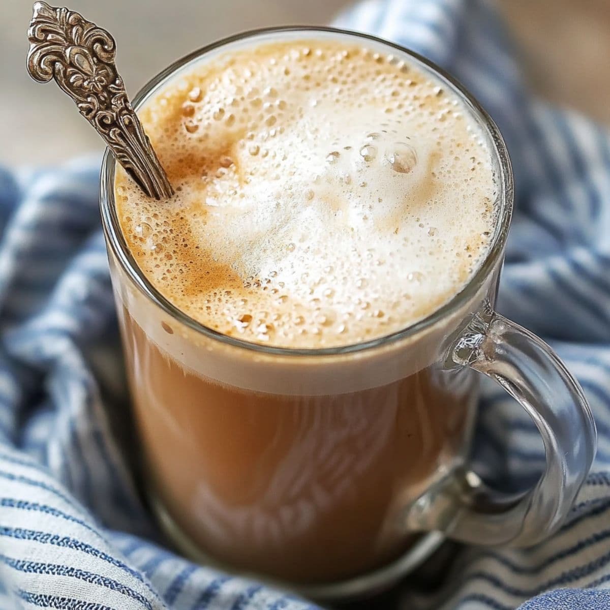 A glass mug of foamy Cuban cortadito coffee with a vintage spoon, wrapped in a blue-striped cloth.