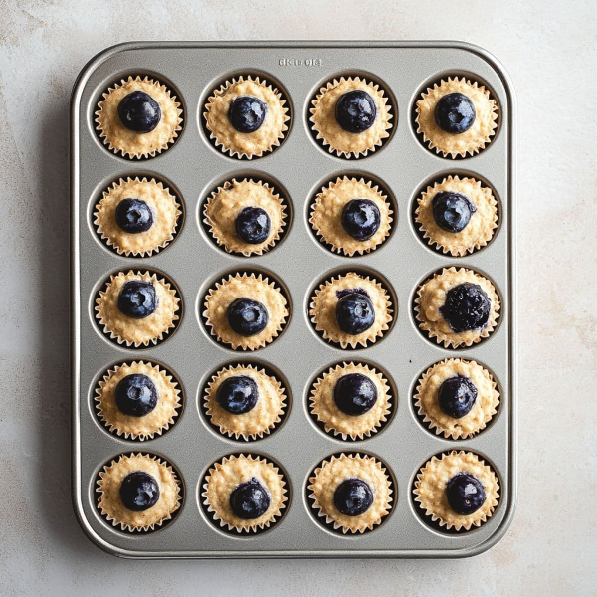 Mini muffin tin filled with raw blueberry muffin batter in paper liners, each topped with a single blueberry, ready to bake.