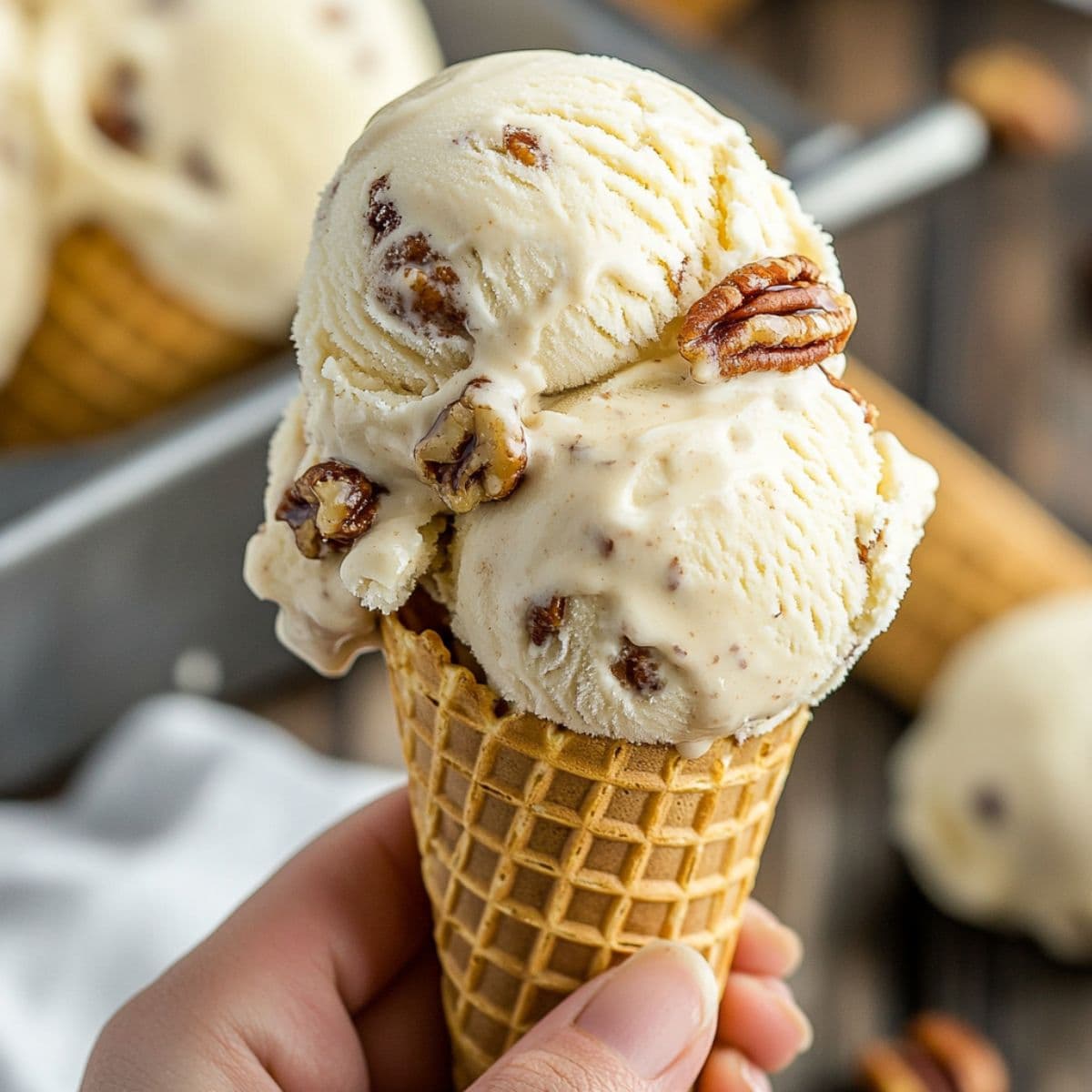 Hand holding a cone of butter pecan ice cream with melting scoops and visible pecan chunks in a warm, rustic setting.