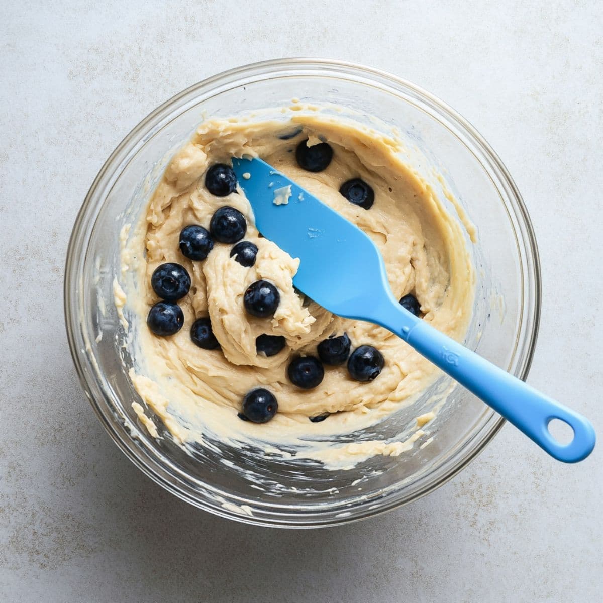 Thick muffin batter with whole blueberries being gently folded using a blue silicone spatula in a glass bowl on a light kitchen surface.