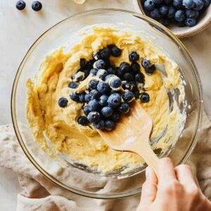 Hand folding blueberries into thick lemon batter with a wooden spatula in a glass bowl.