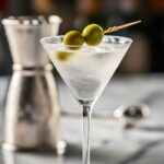 Gin Martini Recipe - Perfect Classic Cocktail in 5 Minutes 13 Chilled gin martini in a cocktail glass with two green olives, on marble with bar tools blurred in the background, a perfect visual for a Gin Martini Recipe.