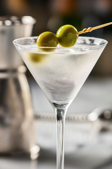 Chilled gin martini in a cocktail glass with two green olives, on marble with bar tools blurred in the background, a perfect visual for a Gin Martini Recipe.