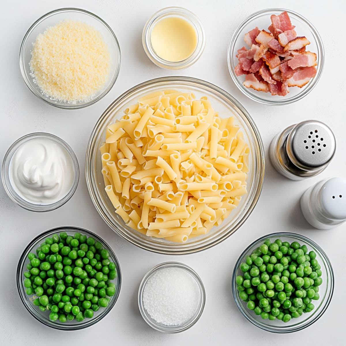 Flat lay of uncooked ditalini pasta, peas, bacon, Parmesan, cream, salt, and pepper in glass bowls on a white surface.