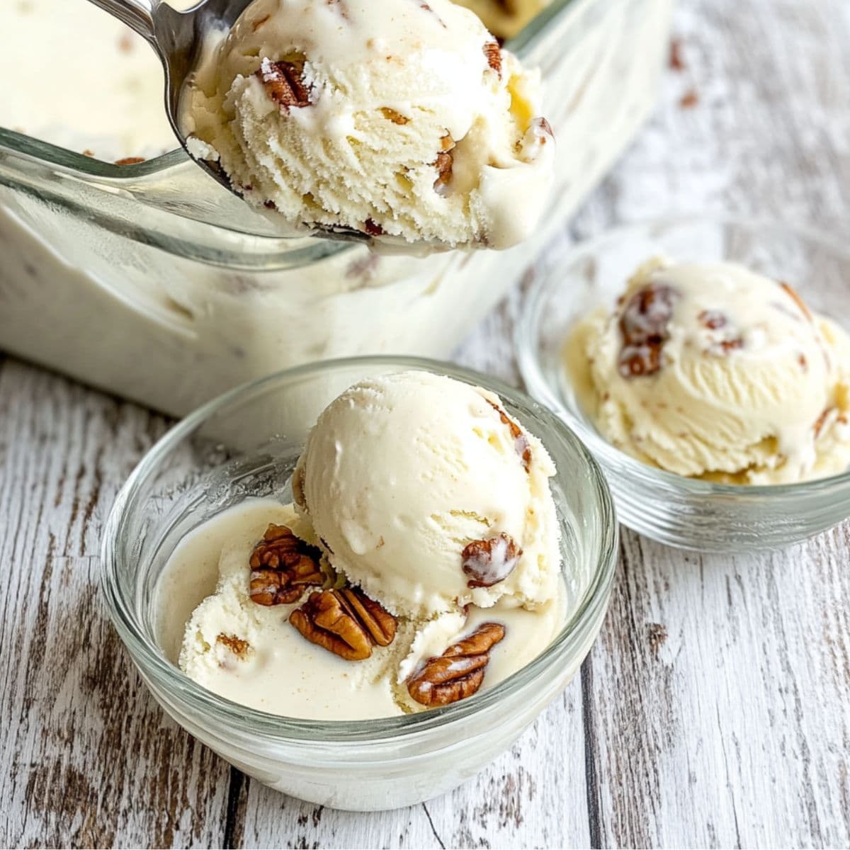 Scoops of butter pecan ice cream served in small glass bowls with pecans on top, with a metal spoon lifting one scoop from a storage container.