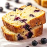 Two slices of lemon blueberry bread stacked on parchment paper with scattered fresh blueberries.