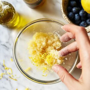 Rubbing lemon zest into cane sugar in a glass bowl with fingers, a key step for enhancing citrus flavor.