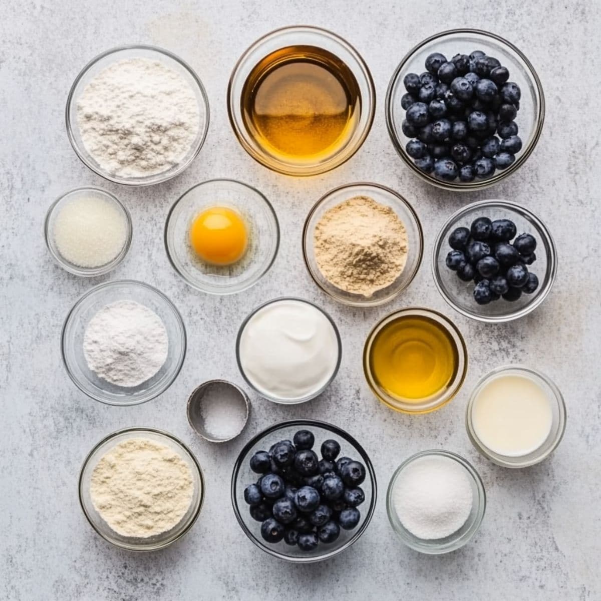 Flat lay photo of measured ingredients for mini blueberry muffins, including blueberries, flour, egg, sugars, sour cream, and lemon zest in small glass bowls on a light gray surface.