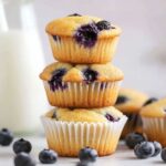 Stack of three mini blueberry muffins with visible blueberries, placed on a white surface with scattered berries and a milk bottle in the background.