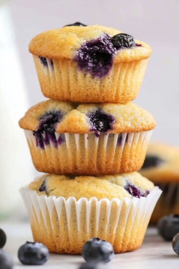 Stack of three mini blueberry muffins with visible blueberries, placed on a white surface with scattered berries and a milk bottle in the background.