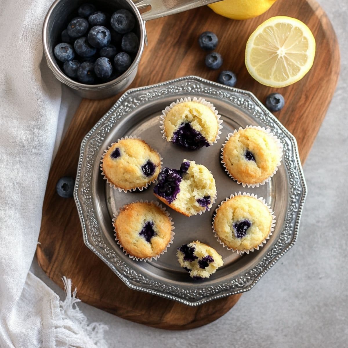 Mini blueberry muffins served on a silver plate with halved muffins, fresh blueberries, and lemon slices on a wooden board.