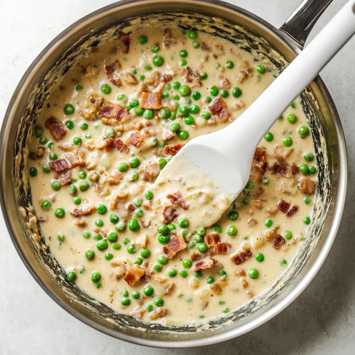 Creamy bacon and pea sauce being stirred in a skillet with a white spatula before adding pasta.
