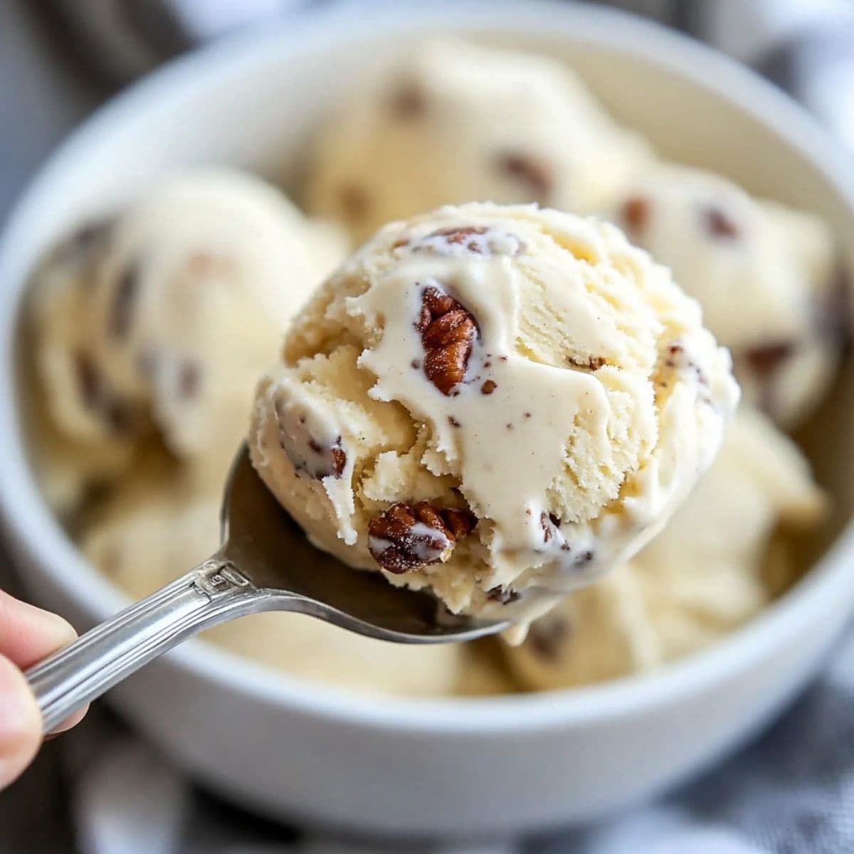 Close-up of a spoon holding a scoop of butter pecan ice cream with roasted pecan pieces, above a white bowl filled with more scoops.