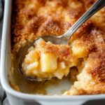 Close-up of pineapple dump cake in a baking dish with golden topping and spoon serving a warm portion.
