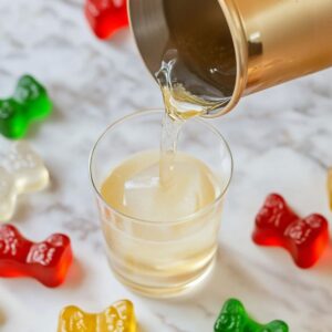 Golden gummy bear cocktail being poured into a glass with ice, surrounded by colorful gummy bears