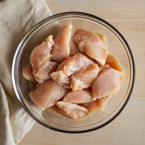 Raw sliced chicken breast in a glass bowl, placed on a wooden surface beside a beige cloth.