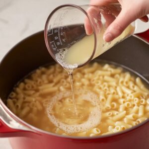 Hand pouring starchy pasta water from a measuring cup back into a pot of cooked ditalini pasta.