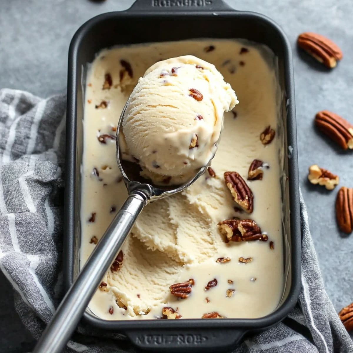 Scooping butter pecan ice cream from a metal loaf pan, showing creamy texture with visible roasted pecans.