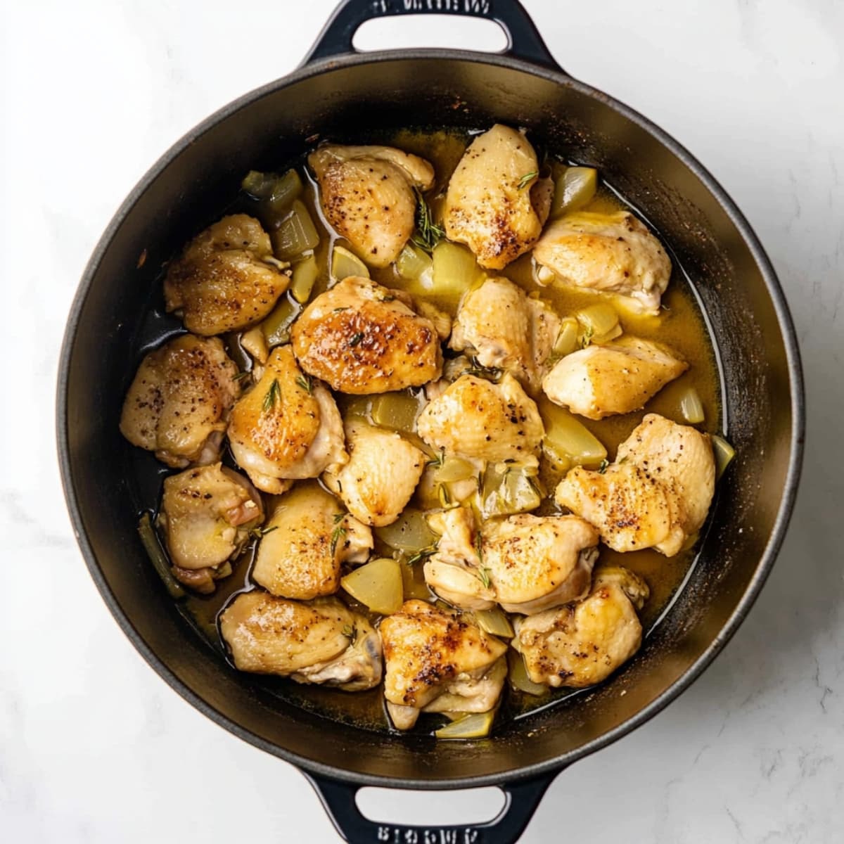 Golden seared chicken simmering in broth with onions inside a black Dutch oven.