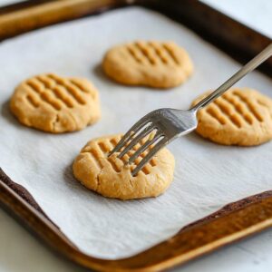 Fork pressing cross-hatch pattern into peanut butter cookie dough on baking sheet.