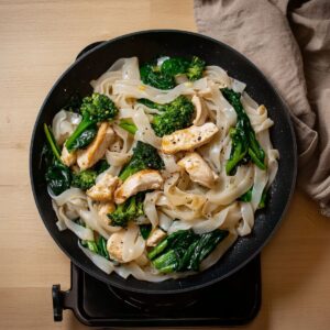 Chicken, flat rice noodles, and greens stir-fried in a non-stick pan over a stovetop.