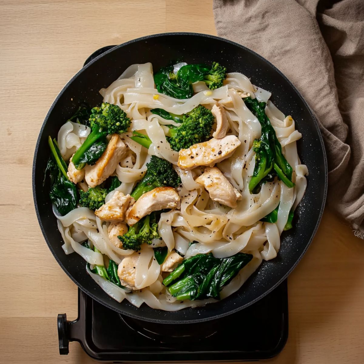 Chicken, flat rice noodles, and greens stir-fried in a non-stick pan over a stovetop.