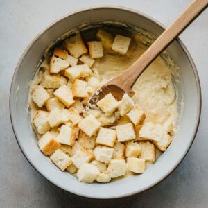 Bread cubes being mixed into pineapple stuffing batter with a wooden spoon, ready to transfer to baking dish