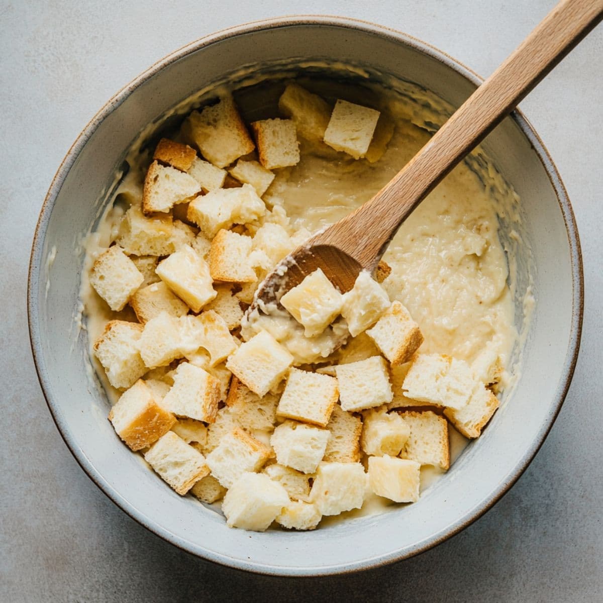 Bread cubes being mixed into pineapple stuffing batter with a wooden spoon, ready to transfer to baking dish