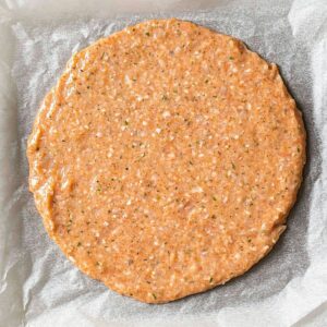 A round, uncooked crust made from ground ingredients, spread evenly on parchment paper, ready to be baked.