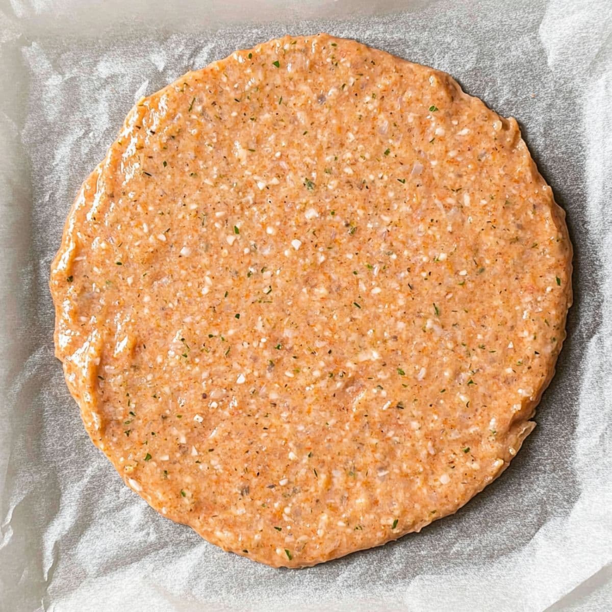 A round, uncooked crust made from ground ingredients, spread evenly on parchment paper, ready to be baked.