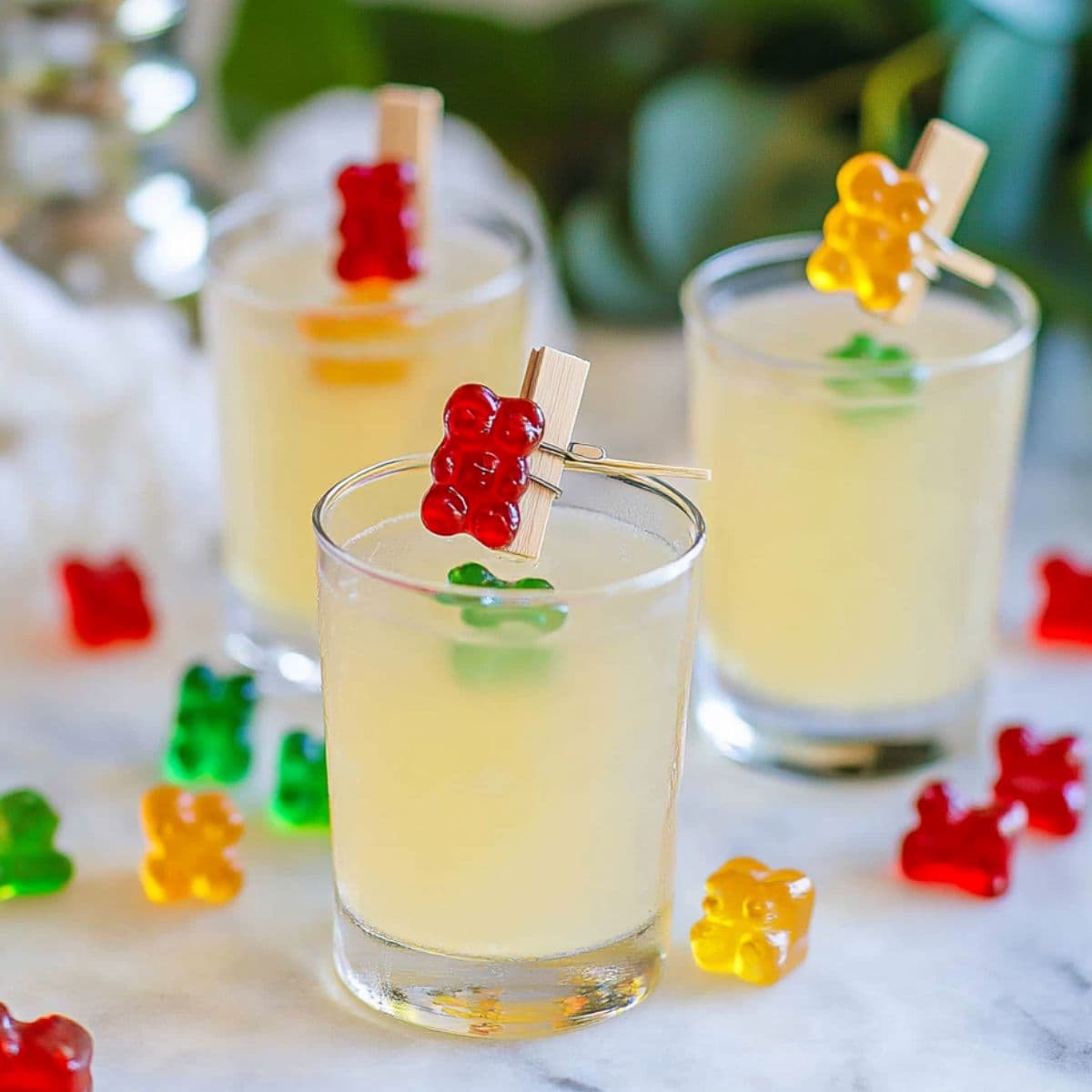Three gummy bear shots with red, yellow, and green gummies clipped to the rims, surrounded by candy on marble