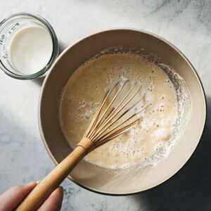 Hand whisking vanilla pudding mix with milk in a beige bowl.