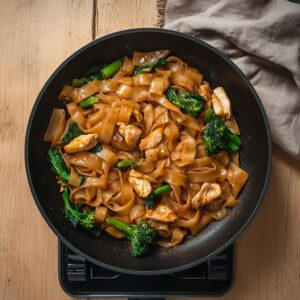Dark soy-coated rice noodles stir-fried with chicken and broccoli in a non-stick pan.