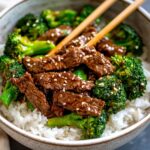 Close-up of beef and broccoli stir fry served over steamed white rice in a ceramic bowl with chopsticks on top.