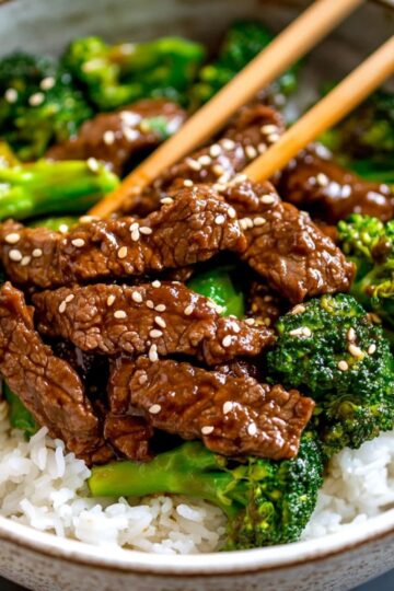 Close-up of beef and broccoli stir fry served over steamed white rice in a ceramic bowl with chopsticks on top.
