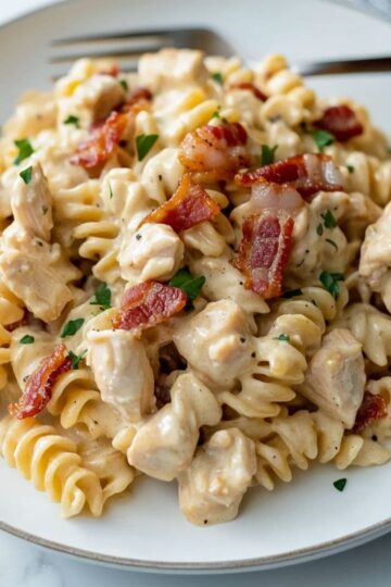 Close-up of creamy chicken bacon ranch pasta with rotini, bacon bits, and chopped parsley served on a white plate with a fork.