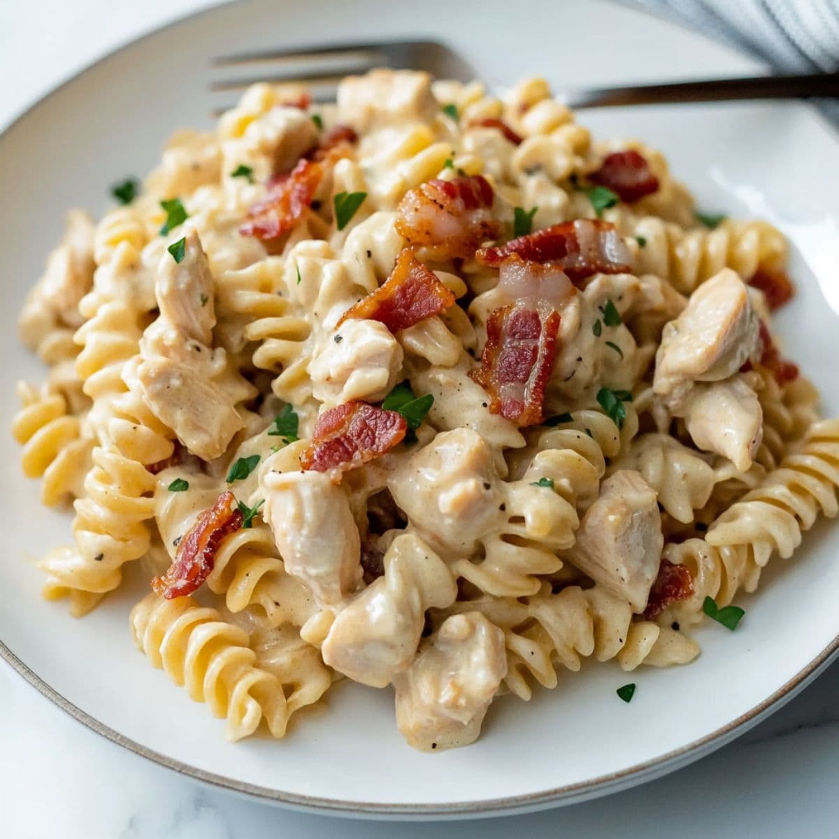 Close-up of creamy chicken bacon ranch pasta with rotini, bacon bits, and chopped parsley served on a white plate with a fork.